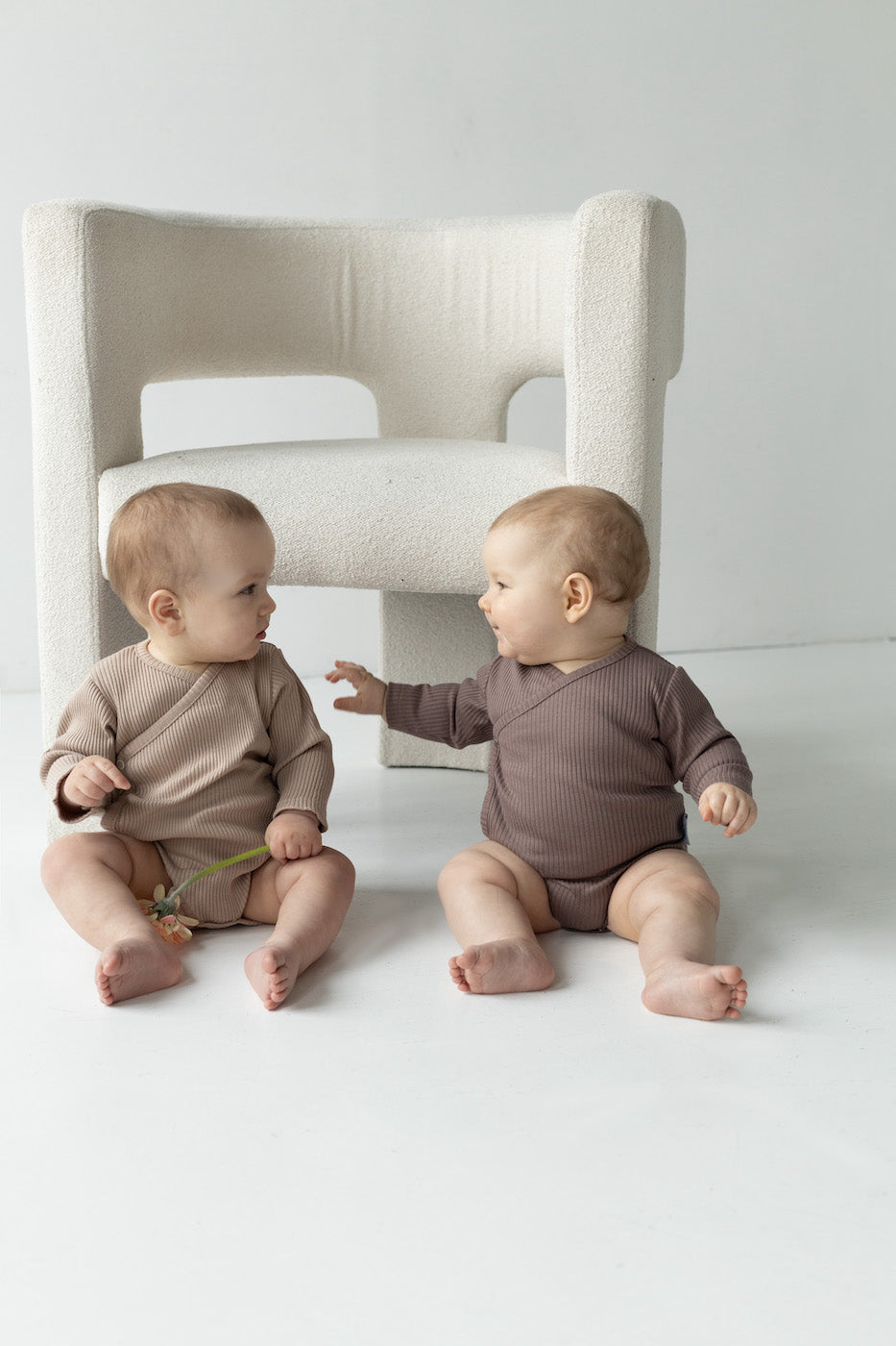 zwei Babys sitzen auf dem Boden vor dem Sessel. Sie habn beige und braue Wickelbody an. 