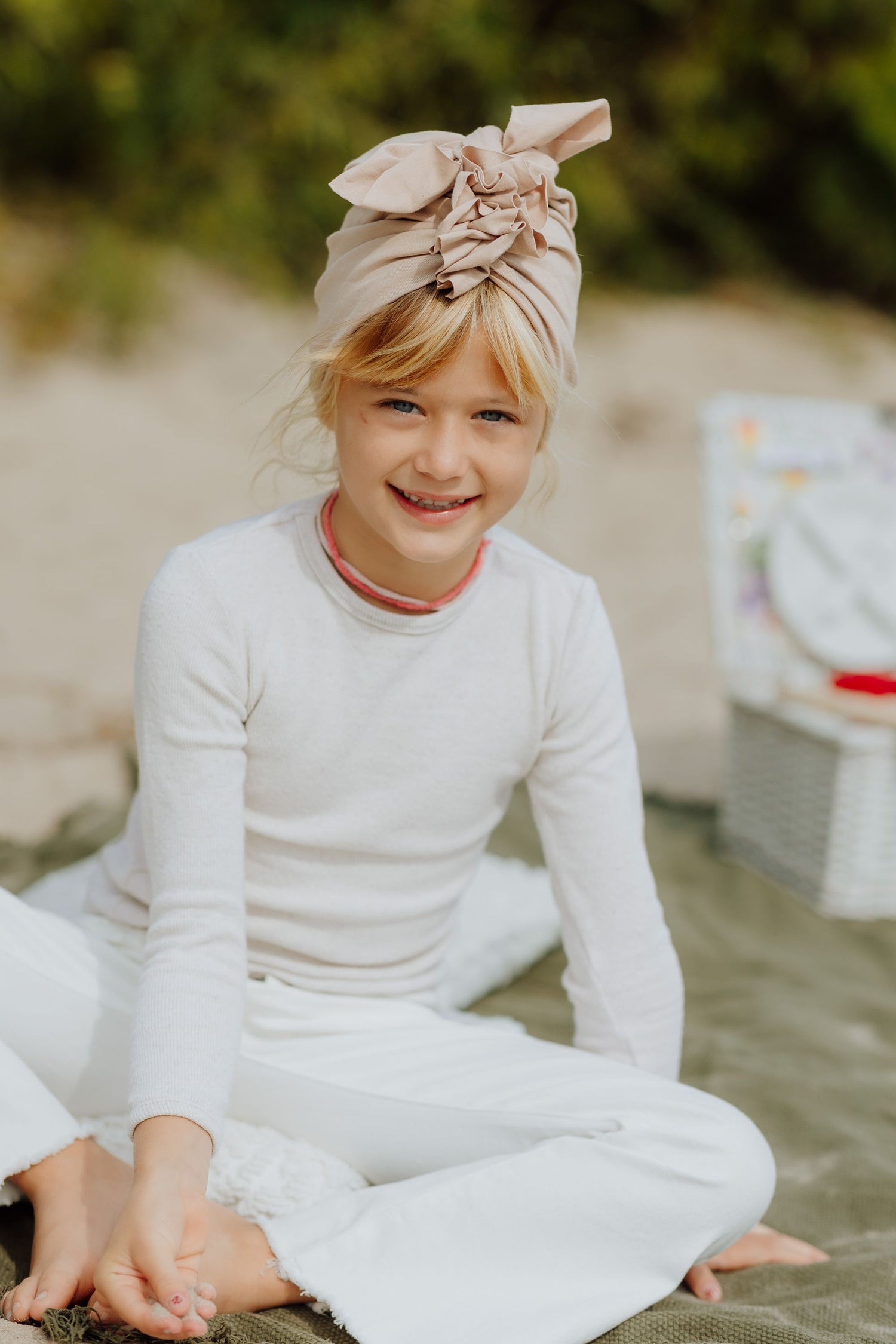 A smiling girl wearing a white shirt and pants and a beige turban sitting on a beach