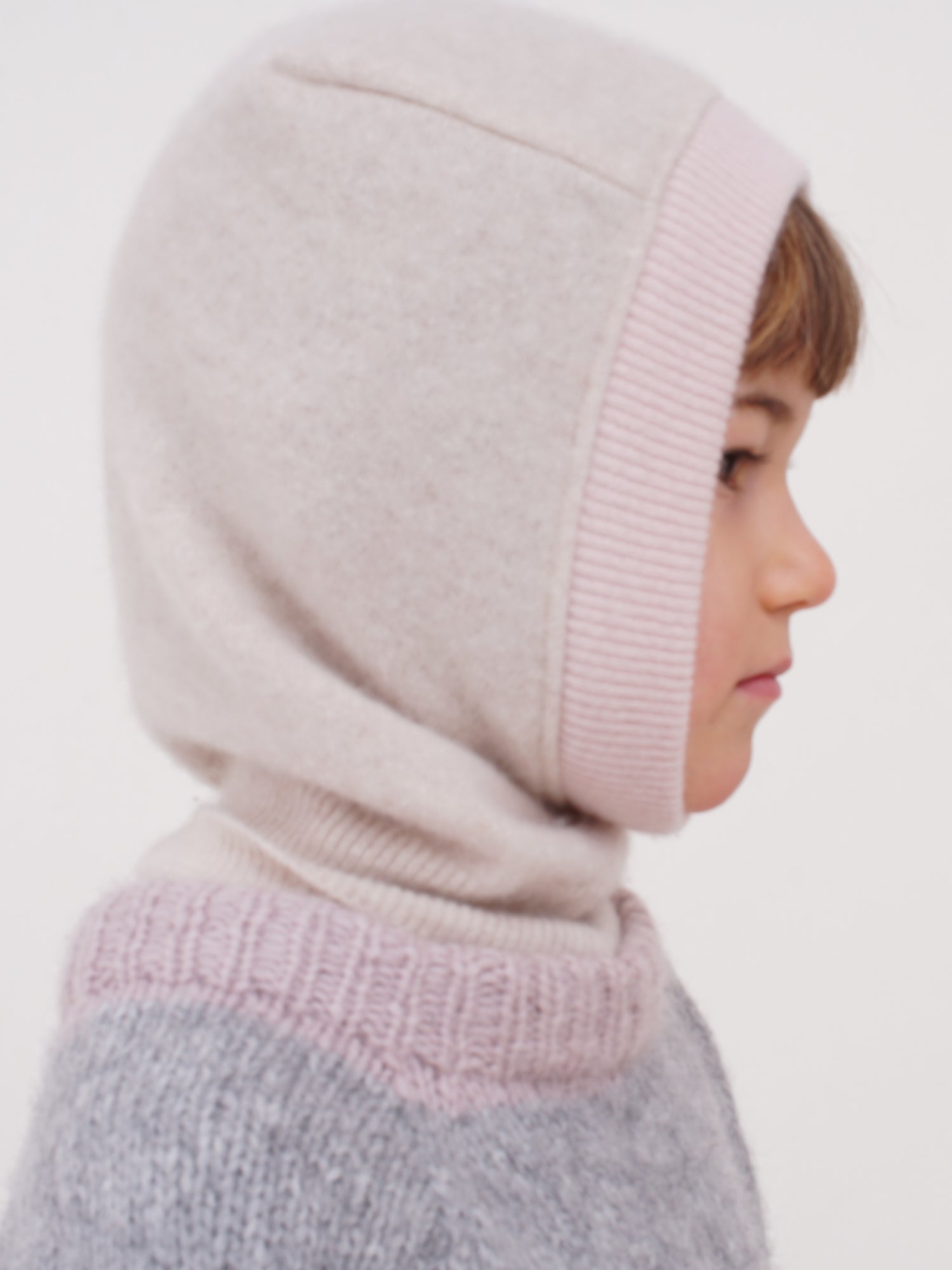 a child wearing a cashmere balaclava in color beige with light pink element and grey sweater looking into the side