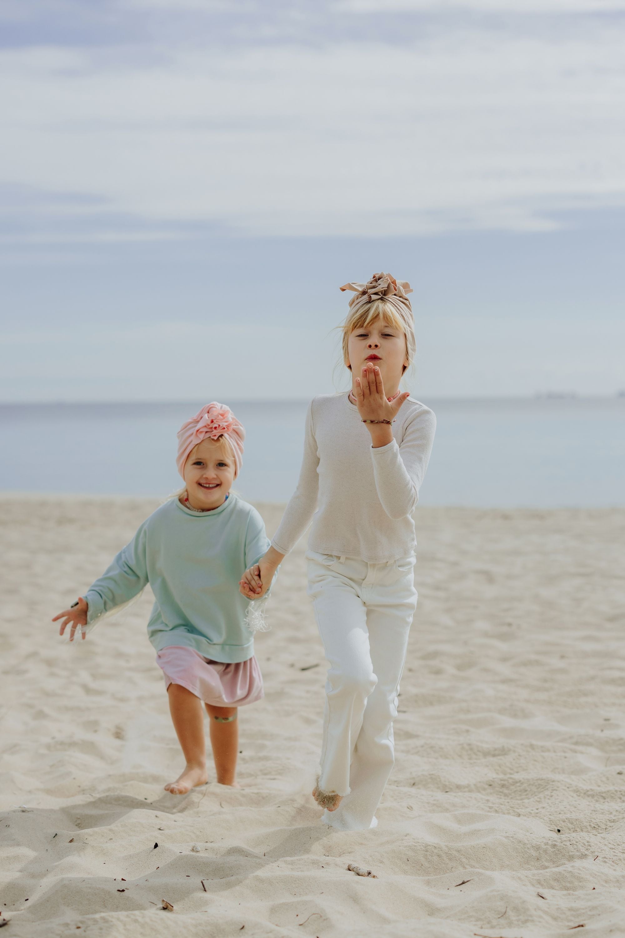 Two running on the beach girls wearing pink and beige turbans. Older girl is wearing white longsleeve and pants and beige turban and is sending a kiss to the camera and younger girl is wearing light blue sweatshirt and pink skirt and pink turban and is smiling. Ocean in the background