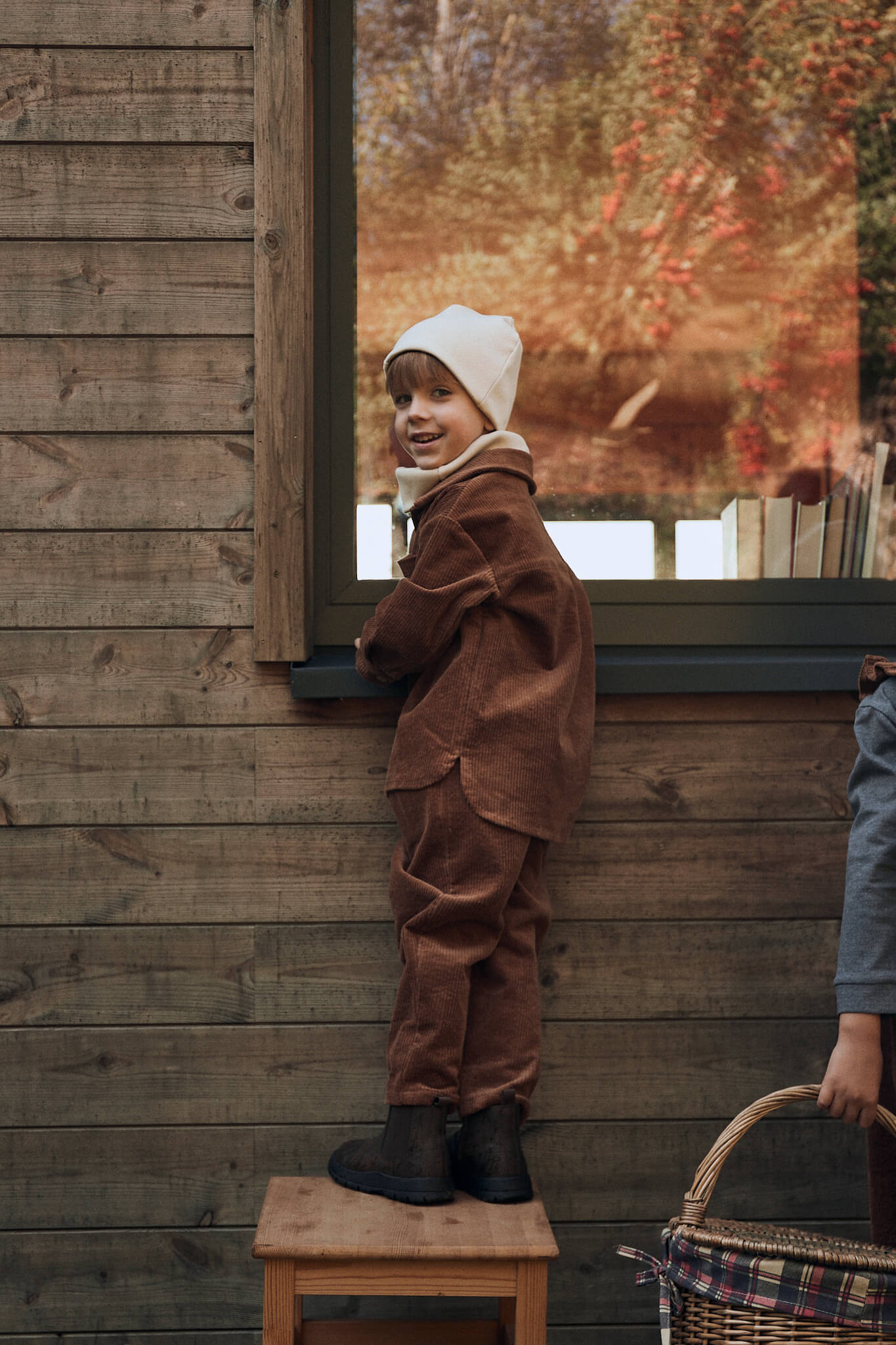 boy wearing brown corduroy set made of jacket and trousers and beige beanie standing at the window