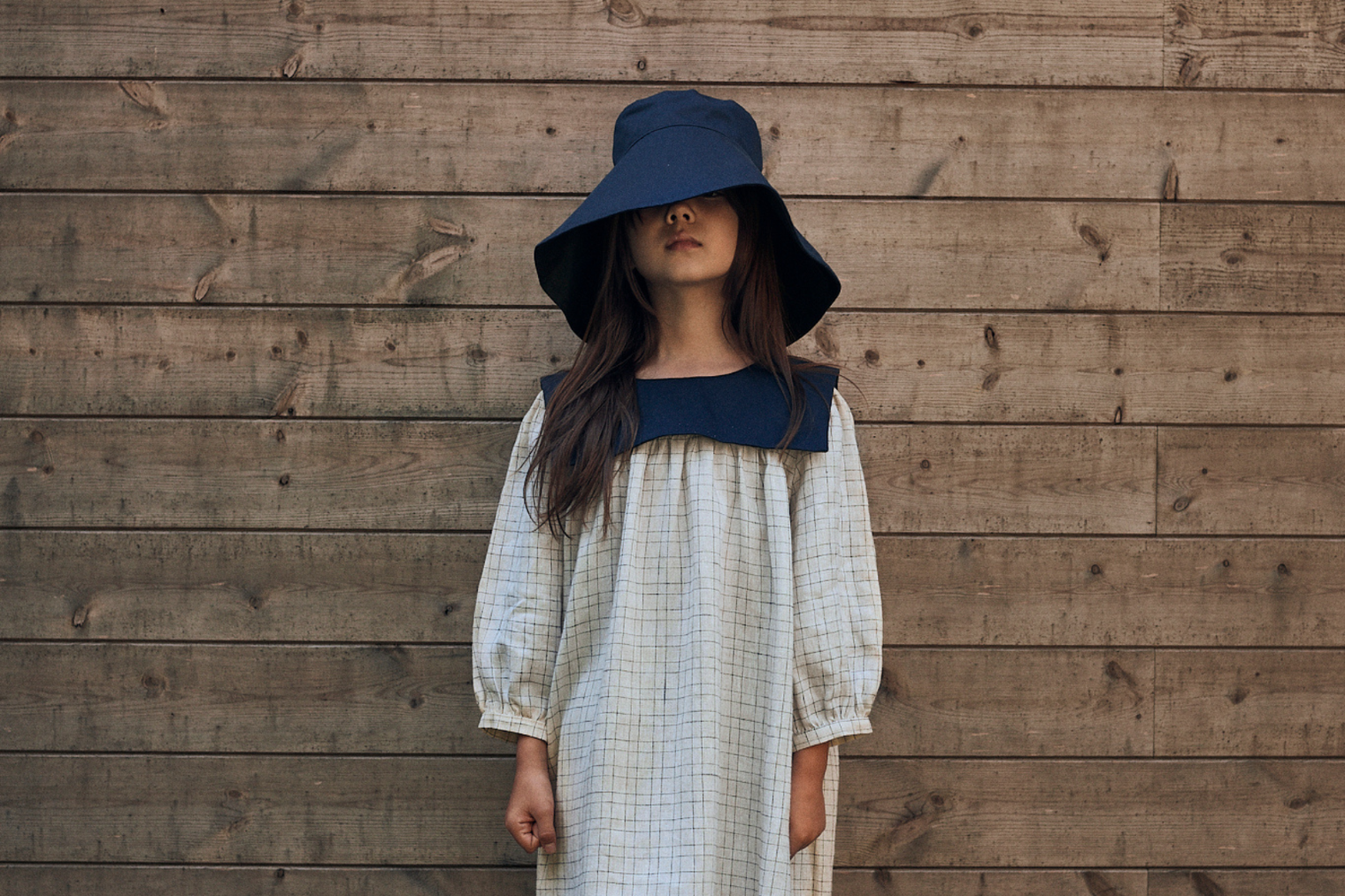 Person wearing a blue hat and white dress against a wooden wall