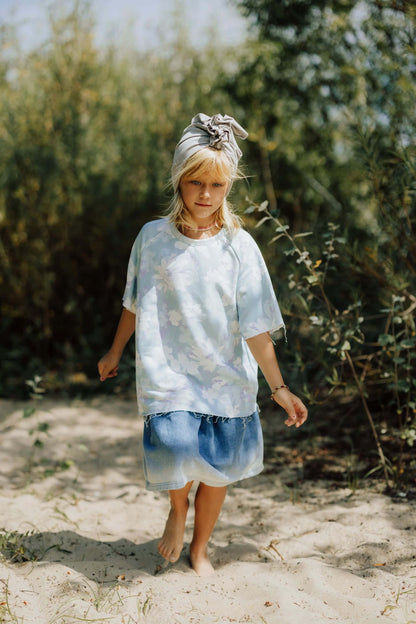 A girl wearing a grey cotton turban and blue shirt and short is walking on the sand. Green trees in the background