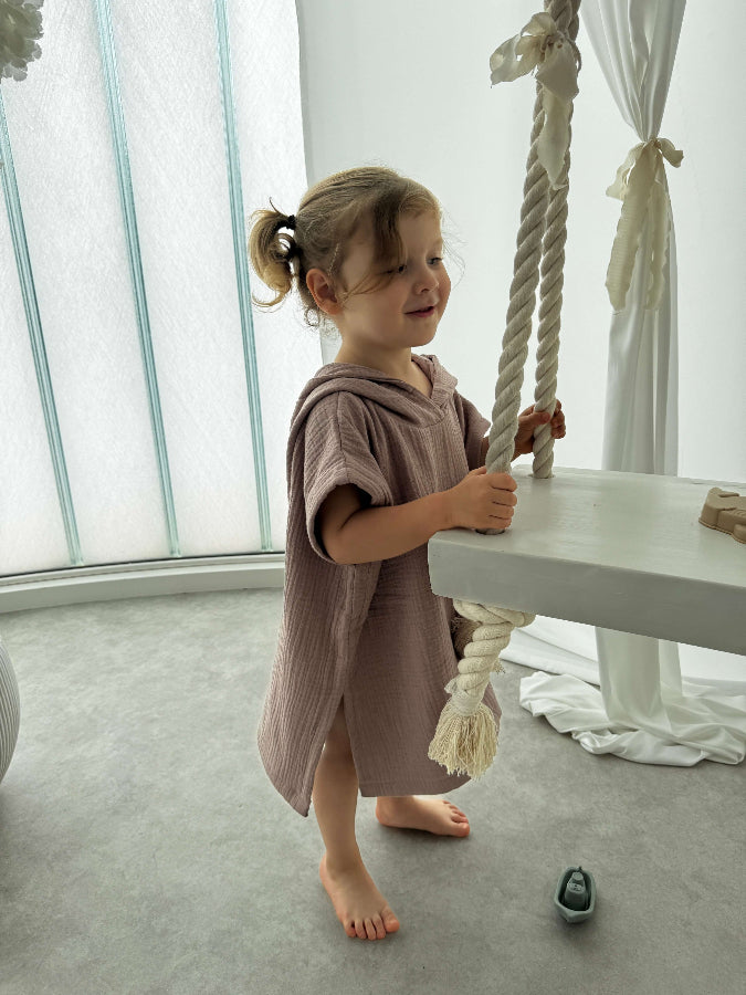 Young girl in a beige poncho standing next to a white table with a rope decoration in a bright room.