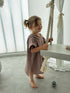 Young girl in a beige poncho standing next to a white table with a rope decoration in a bright room.