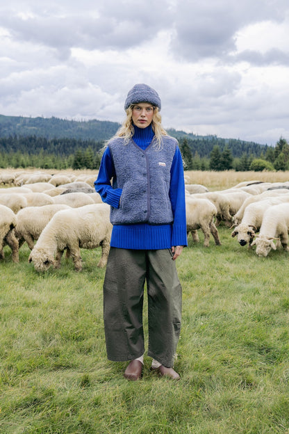 A model stands in a grassy field with sheep in the background, wearing a blue ribbed turtleneck sweater, a gray fleece vest, olive green wide-leg pants, and a gray knit beanie.