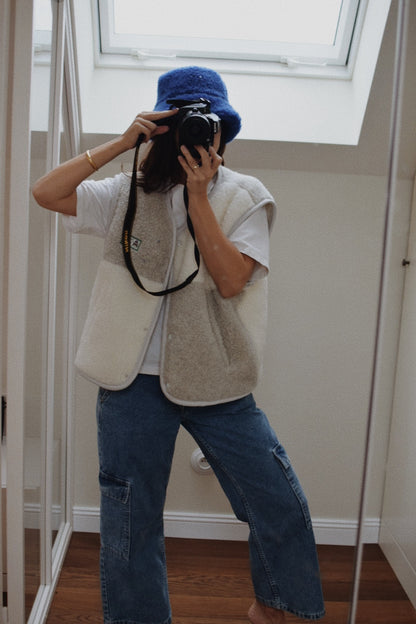Person taking a mirror selfie wearing a blue knit hat, white vest, and blue jeans.