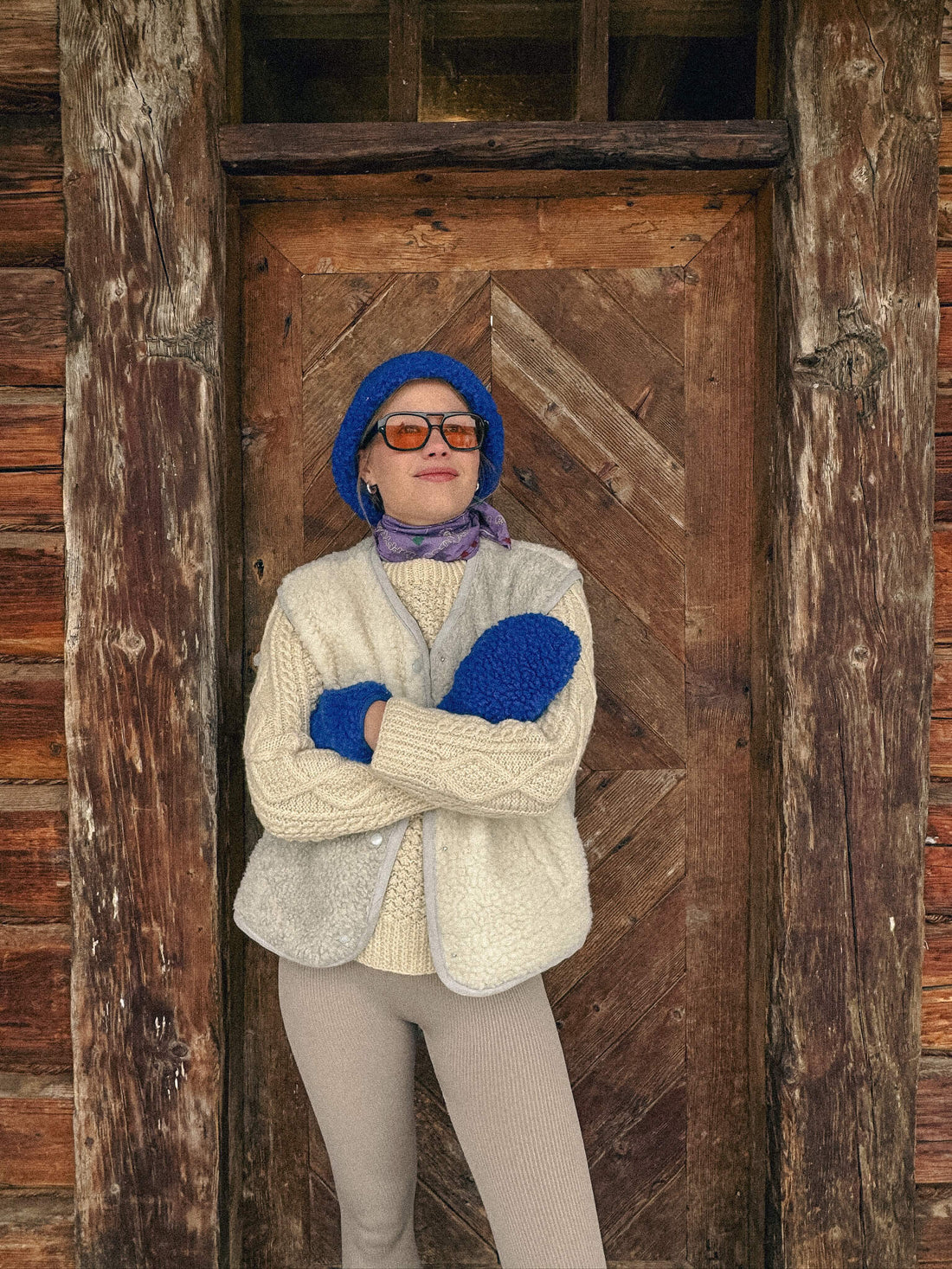 Person wearing a blue hat, gloves, and sunglasses standing in front of a wooden door.