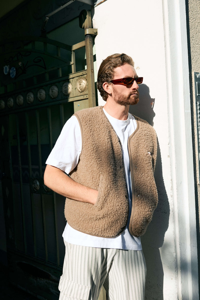 A man wearing a beige sherpa fleece vest over a white t-shirt and striped trousers, standing outdoors against a white wall in bright sunlight.
