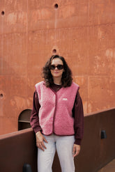 Person wearing a pink vest and sunglasses standing against a rust-colored wall.