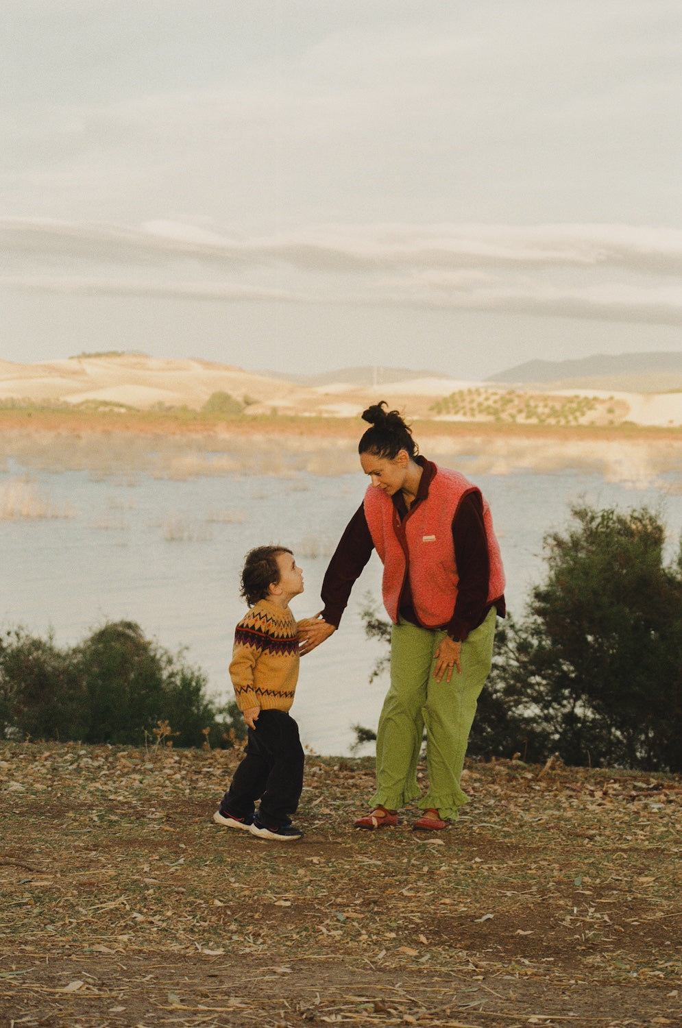 Woman and child holding hands by a lake with mountains in the background
