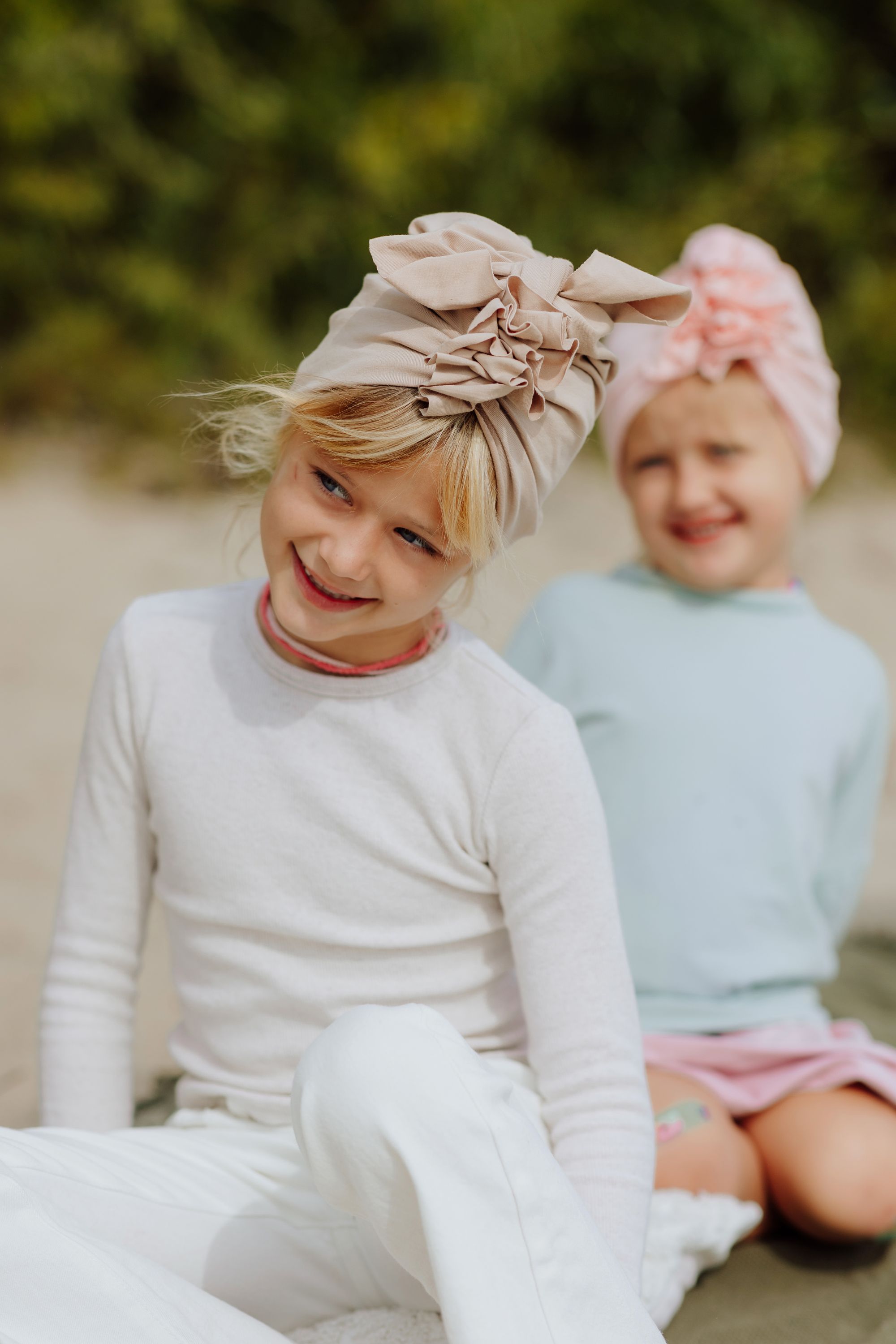 Two smiling girls seating on the beach and wearing pink and beige turbans. Older girl is wearing white longsleeve and pants and beige turban and younger girl is wearing light blue sweatshirt and pink skirt and pink turban and is smiling. Forest in the background