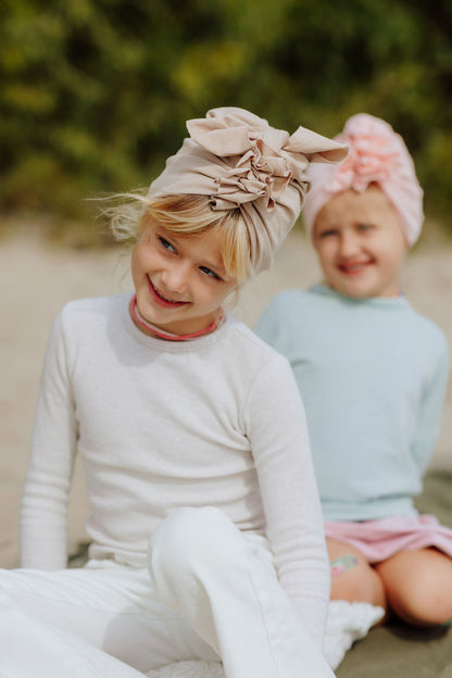 Two smiling girls seating on the beach and wearing pink and beige turbans. Older girl is wearing white longsleeve and pants and beige turban and younger girl is wearing light blue sweatshirt and pink skirt and pink turban and is smiling. Forest in the background