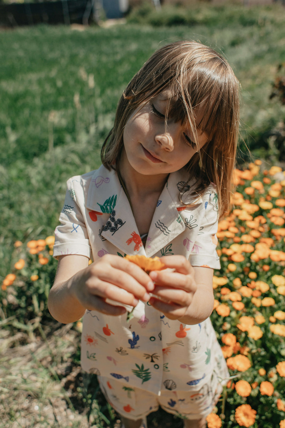 A child standing in front of a flower bed, wearing a linen shirt with a safari print, holding a flower.
