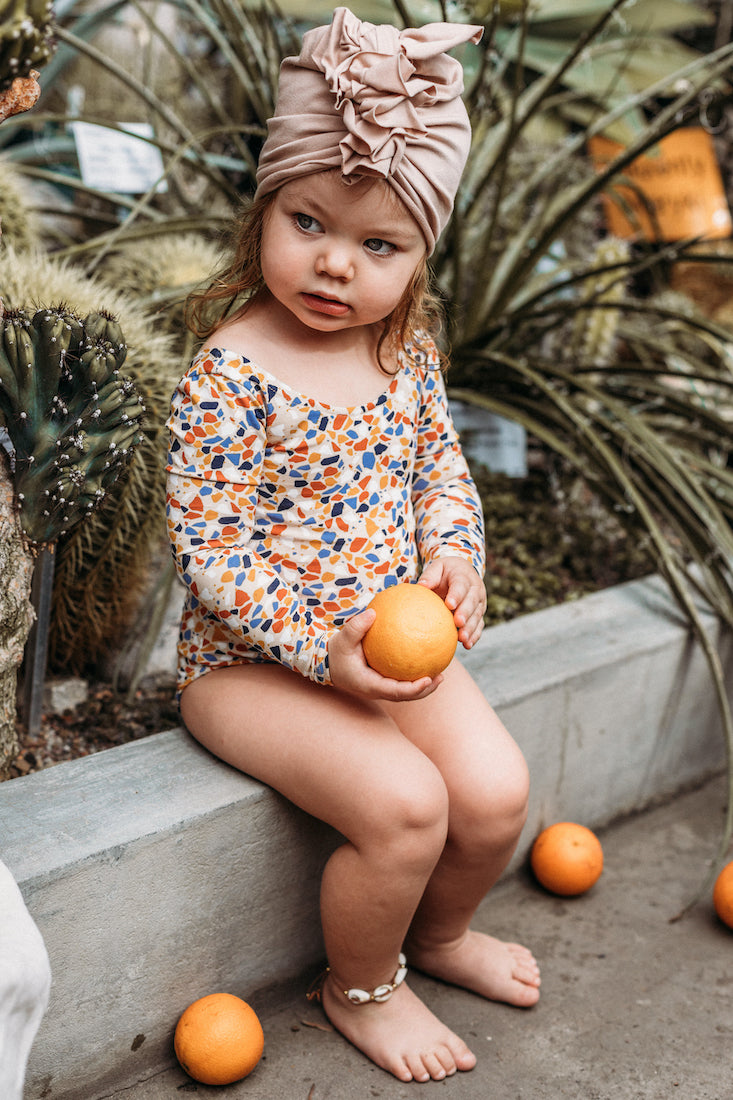 Ein sitzende Mädchen angezogen in bunten Swimmanzug  und beige Mütze Turbane hält ein Orange. 