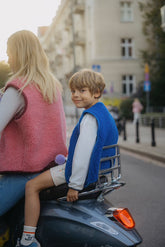 A child sitting on the back of a motorcycle wearing a blue merino wool vest with a smiling expression, accompanied by an adult in a pink vest.