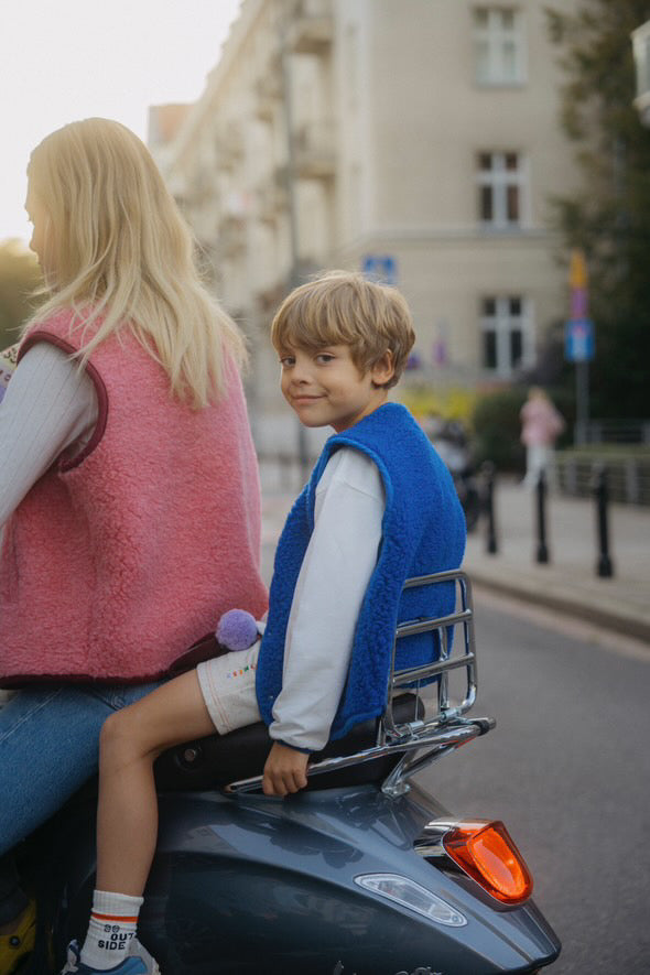 Child sitting on the back of a motorcycle wearing a blue merino wool vest with a smiling expression