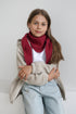 A young girl with long light brown hair is wearing a thick, deep red knit scarf, a beige zip-up cardigan, a white t-shirt, and light blue jeans, sitting against a plain light gray wall.