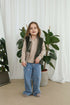 A young girl stands in a bright room wearing a beige long-sleeve top, a dark olive green vest, and light blue wide-leg jeans with small pink embroidered details. She is positioned in front of white curtains and potted green plants.