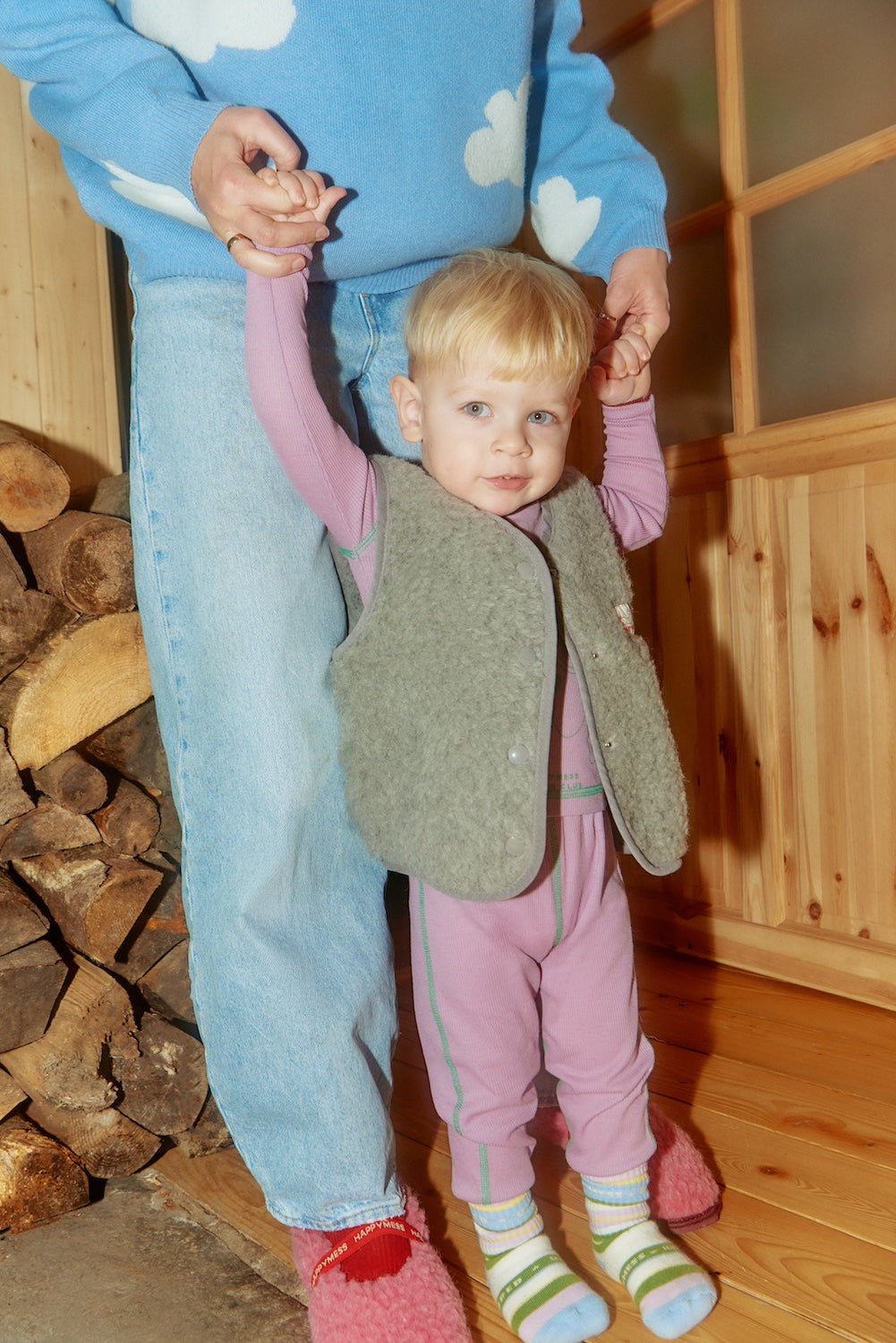 Child in pink outfit with grey vests with an adult in blue outfit holding hands indoors.