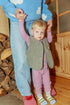 Child in pink outfit with grey vests with an adult in blue outfit holding hands indoors.