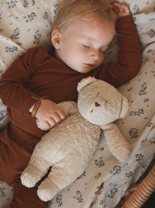 Ein schlafendes Baby in braunem Body und Leggins aus Merinowolle und Seide. Das Baby haltet ein Bär. 