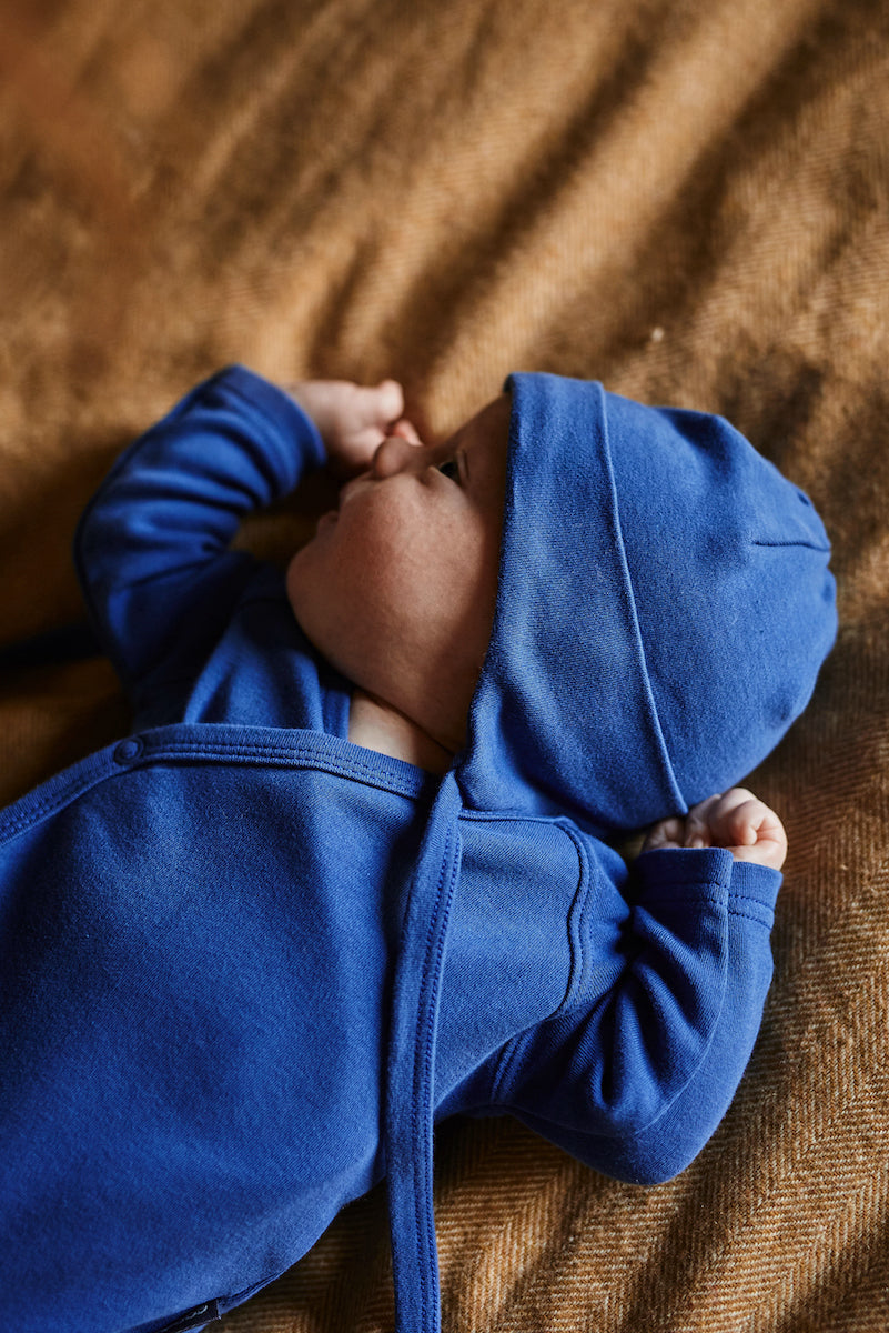 a small baby lying on his back on a brown blanket wearing a cobalt blue bonnet and long sleeve wrap style bodysuit