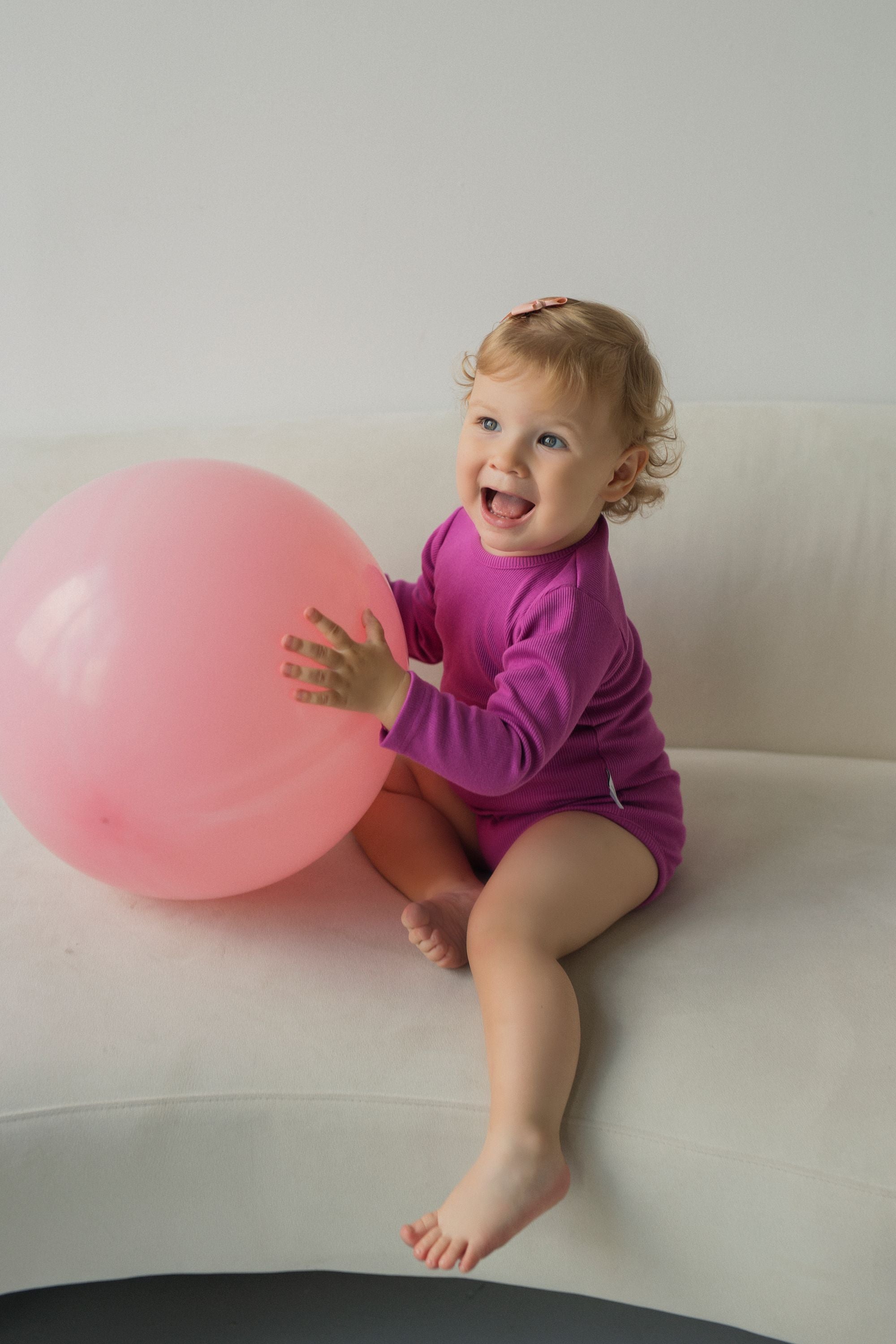 Girl wearing pink bodysuit holding a baloon and sitting on a couch