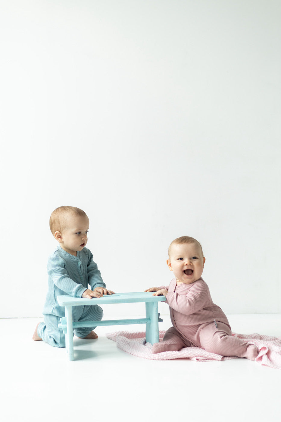 2 babies wearing baby rompers, one in blue and one in pink, smiling and sitting on the blanket