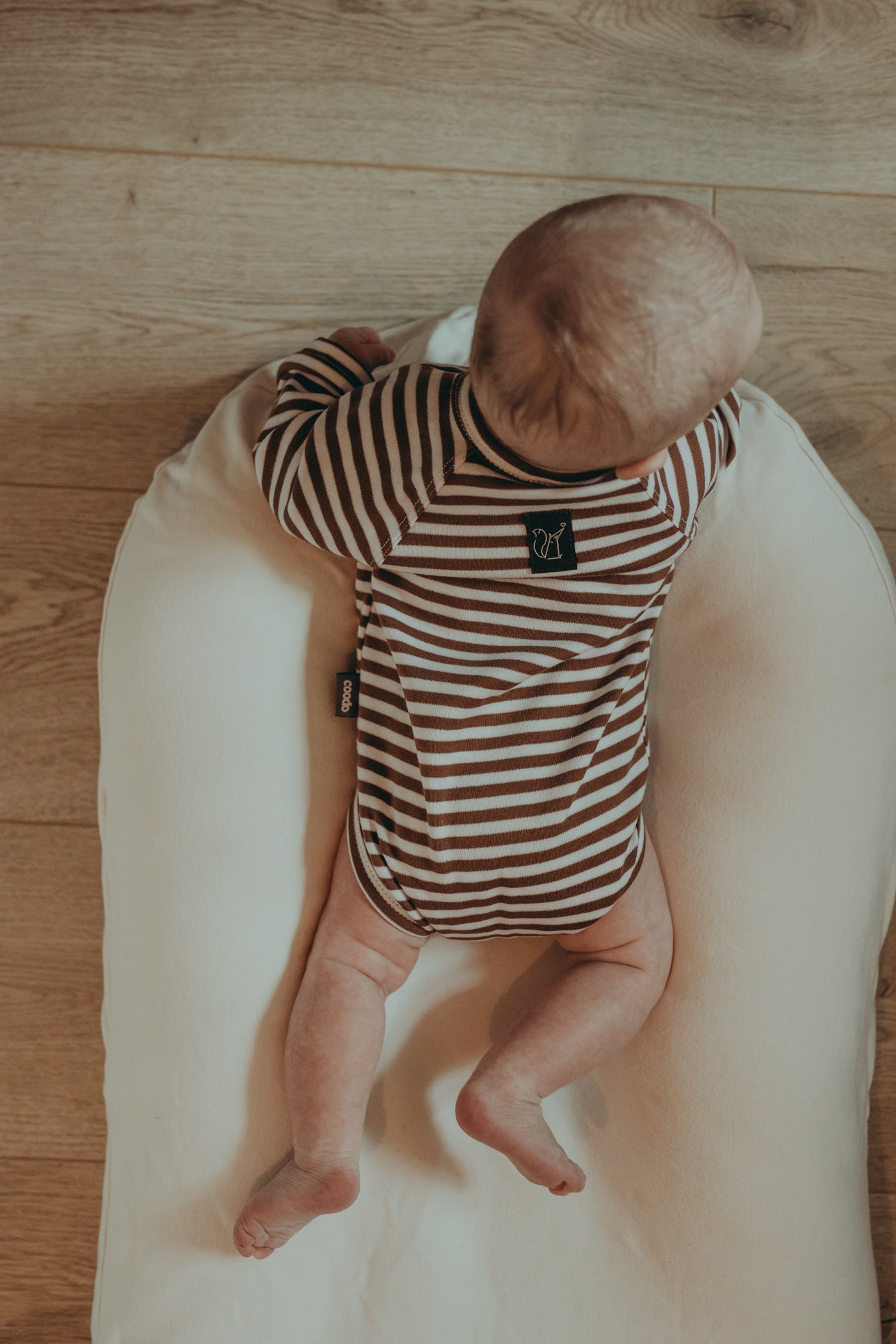 A small baby lying on a belly on a white cocoon wearing a wrap style ecru bodysuit with brown stripes. Cocoon is placed on a wooden floor