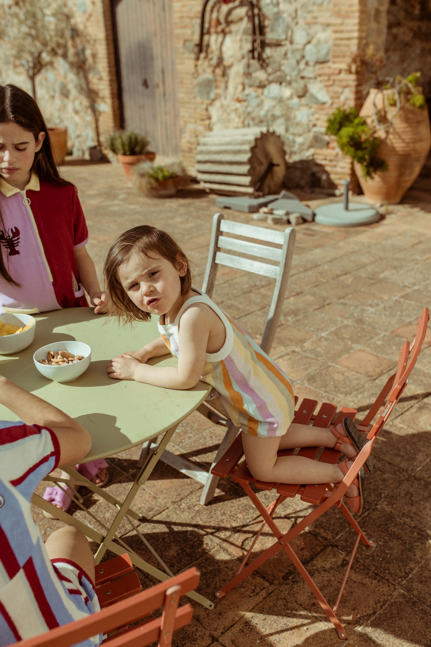 A child sitting at an outdoor table wearing a pink and white striped romper with a palm tree detail, accompanied by an older girl.
