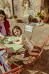 A child sitting at an outdoor table wearing a pink and white striped romper with a palm tree detail, accompanied by an older girl.