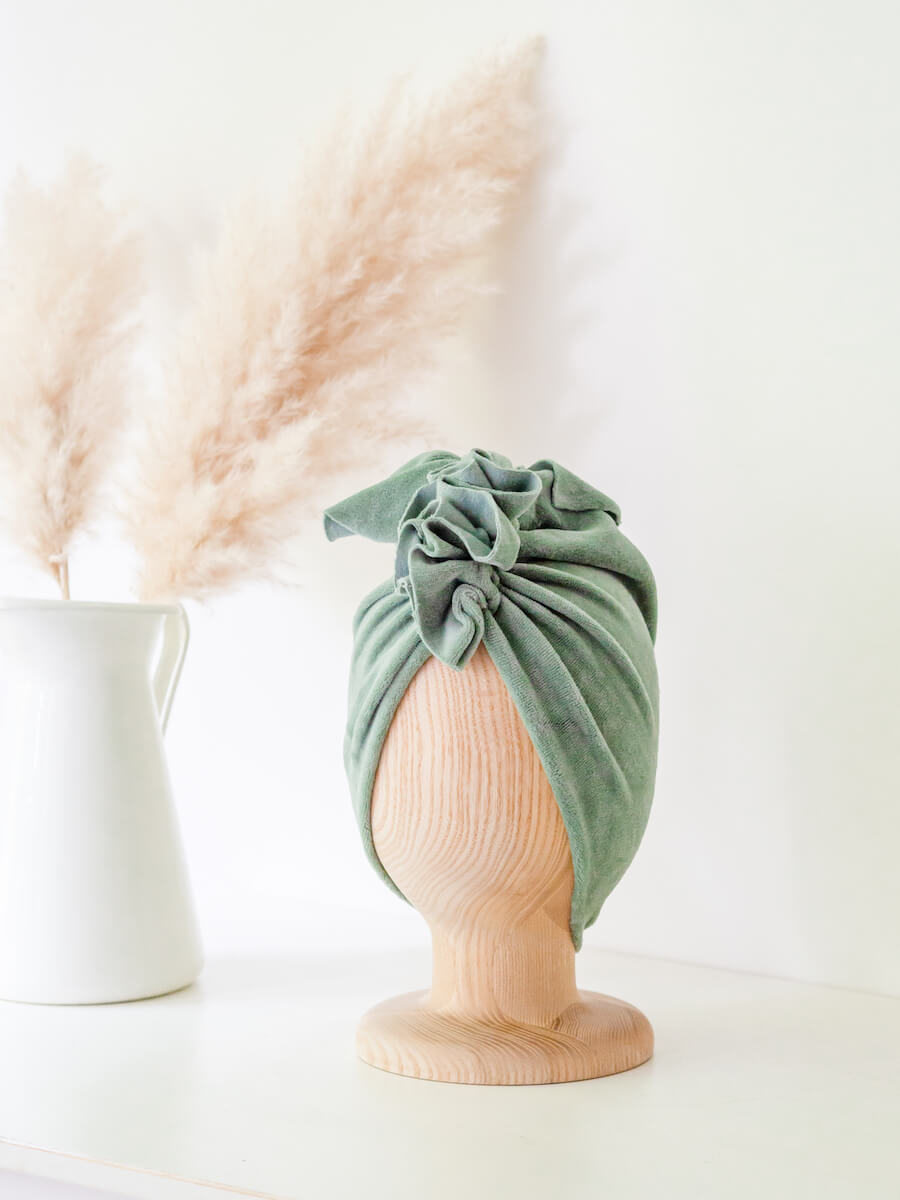 baby turban in color dusty mint on a wooden head mannequin with white vase in the background