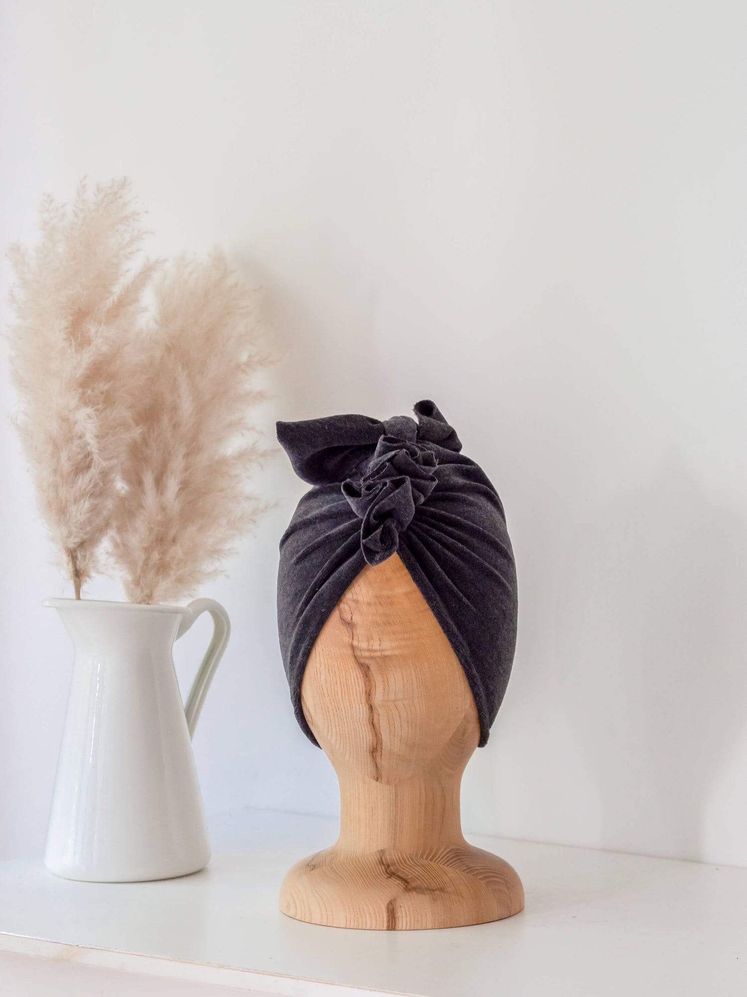 baby turban in color dark grey on a wooden head mannequin with white vase in the background