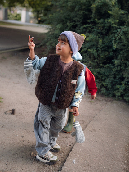 A young child wearing a brown fuzzy vest, light blue cardigan, gray striped pants, and a lavender knit beanie stands outdoors on a paved path near green bushes, looking upward and gesturing with one hand.