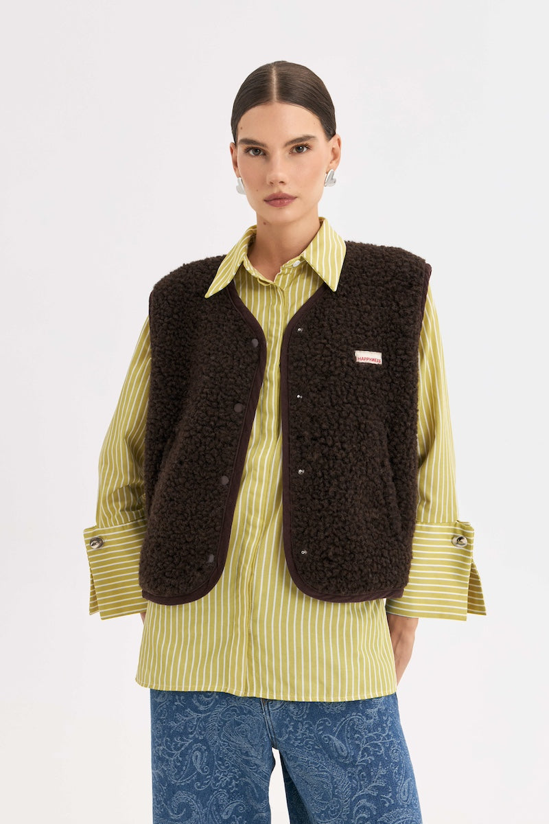 A model wearing a dark brown sherpa fleece button-up vest over a yellow and white striped shirt, paired with blue patterned trousers, against a plain light gray background.