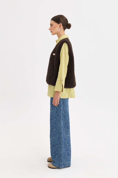 A model shown in profile wearing a dark brown textured vest over a yellow and white striped shirt, paired with wide-leg blue jeans and brown shoes, against a plain white studio background.