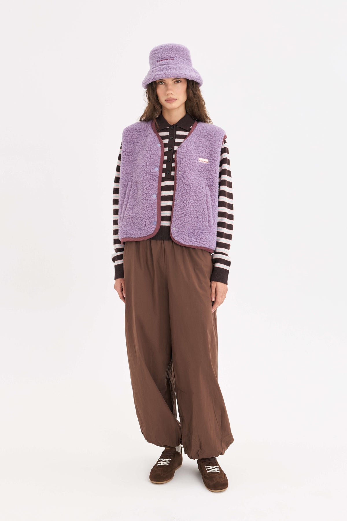 Person wearing brown pants, a lilac cloud merino wool vest over a longsleeve on a white background. 