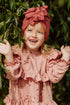 A smiling girl wearing a pink dress with little hearts and dusty red turban on the backdrop from green trees